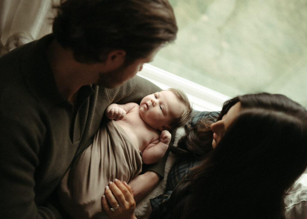Mom and dad looking down at their swaddled newborn baby relaxing on the couch in their northern kentucky home
