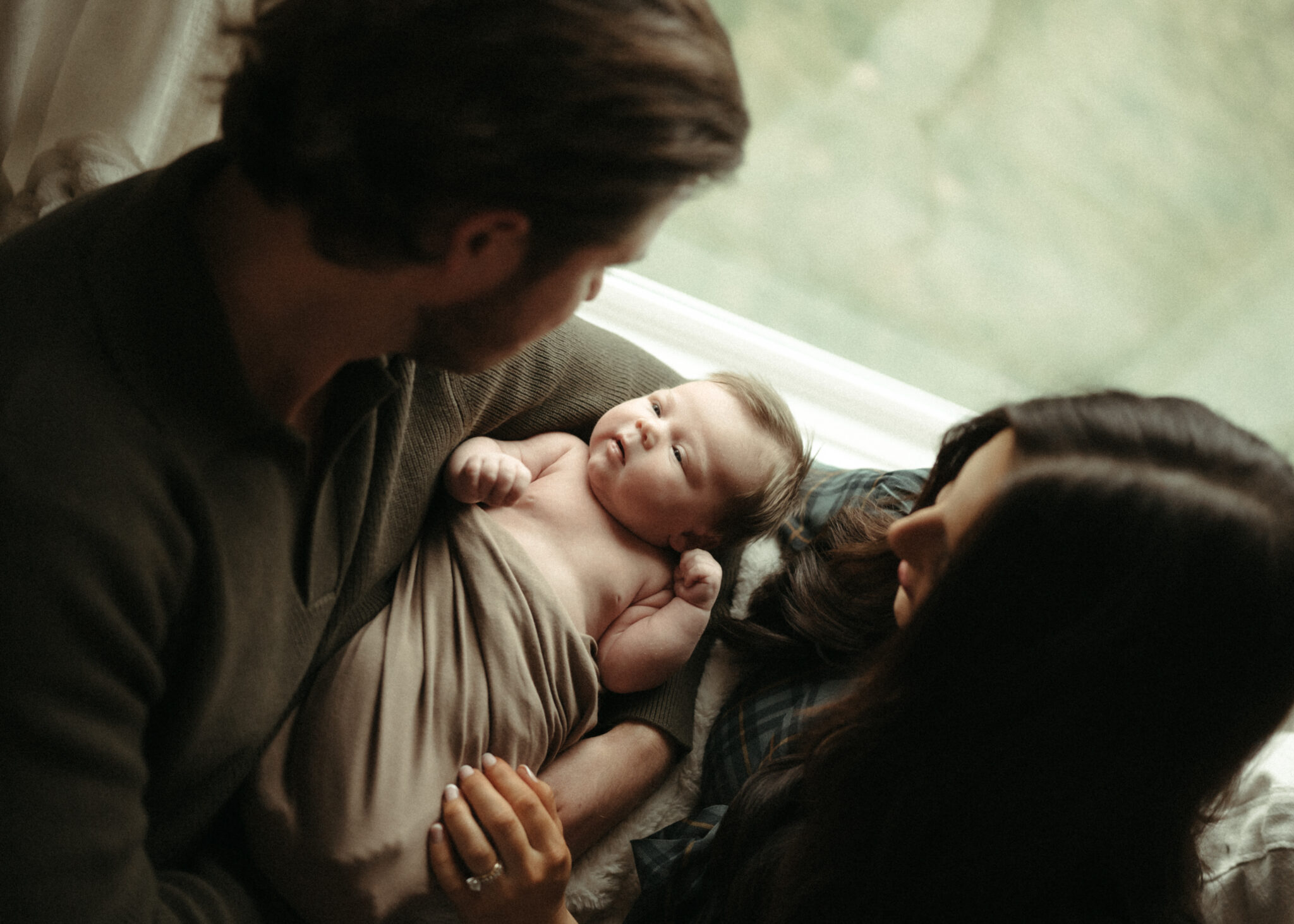 Mom and dad looking down at their swaddled newborn baby relaxing on the couch in their northern kentucky home