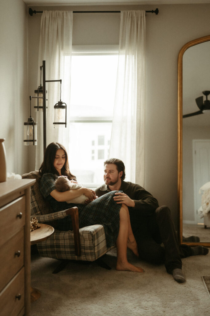 mom holding newborn baby in front of window with dad sitting on the floor during their lifestyle in home newborn photography session