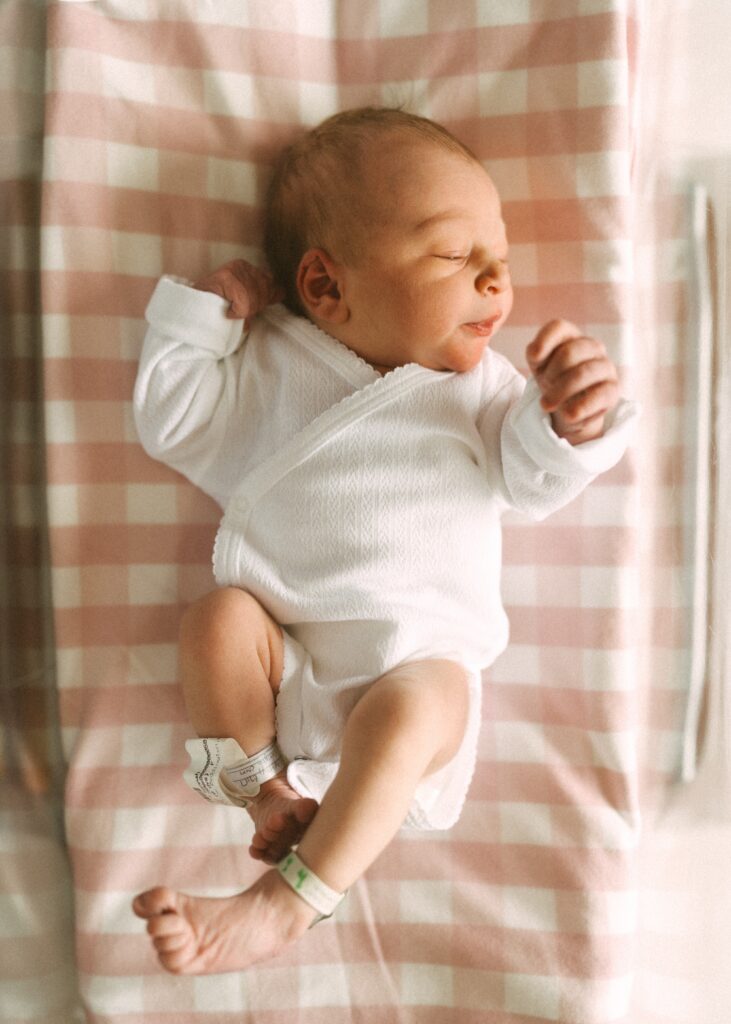 newborn girl laying on pink buffalo check blanket in hospital bassinet