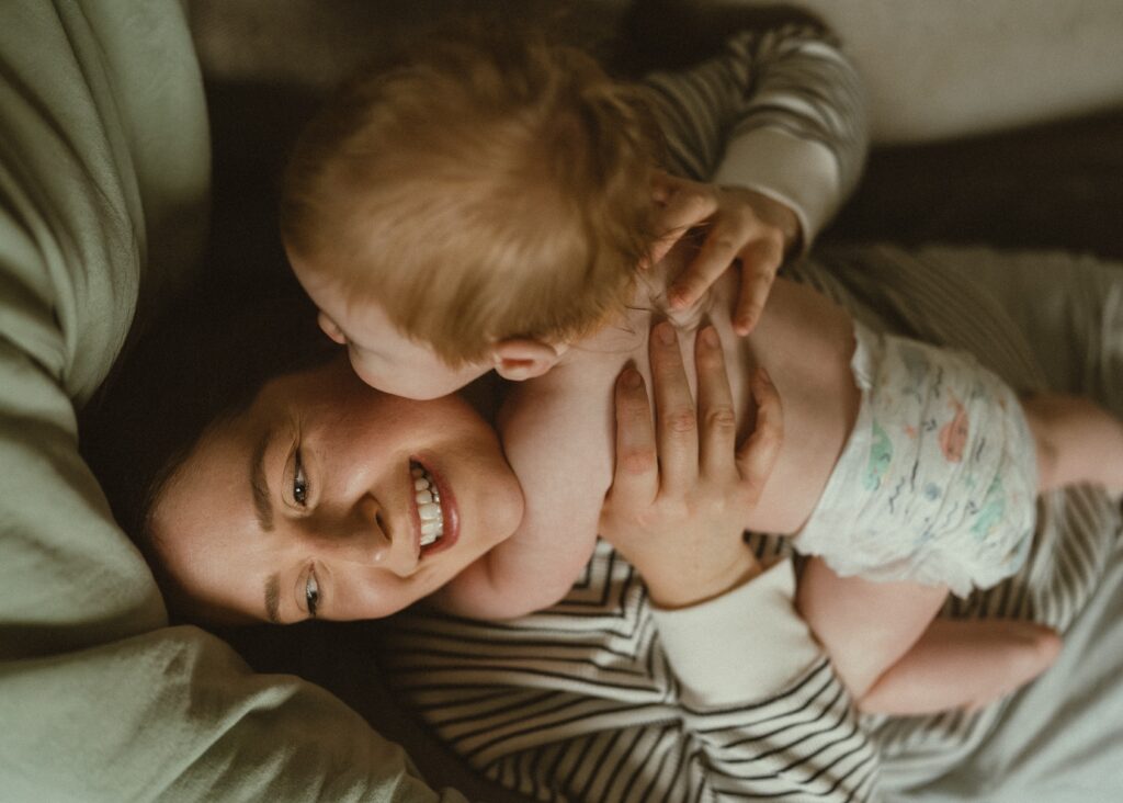 Toddler boy wearing a diaper laying on his mother's chest while he kisses her and she laughs