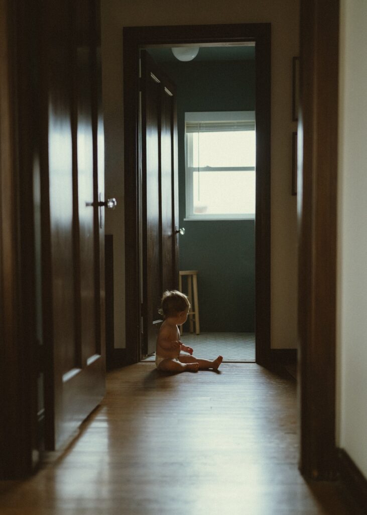 Toddler boy wearing a diaper sitting in a dimly lit hallway during his in home milestone lifestyle session