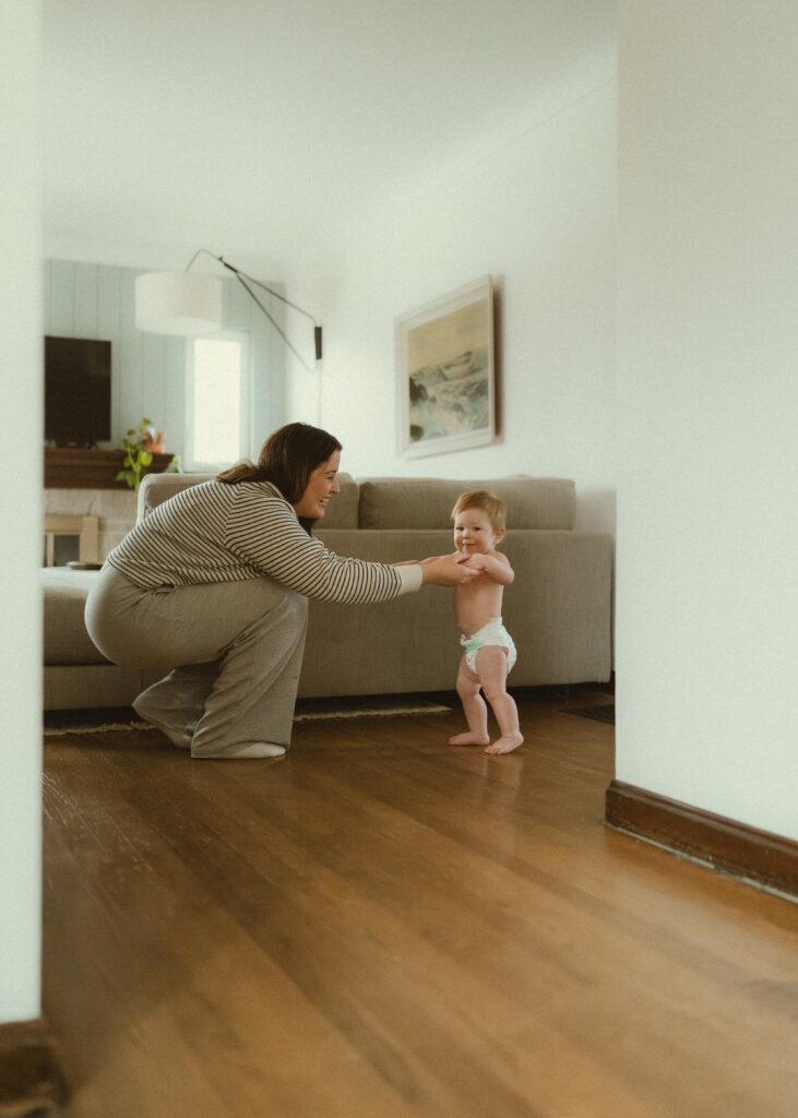 Toddler boy looking and smiling at camera while his mom assists him taking his first steps in their neutral living room in their Cincinnati home