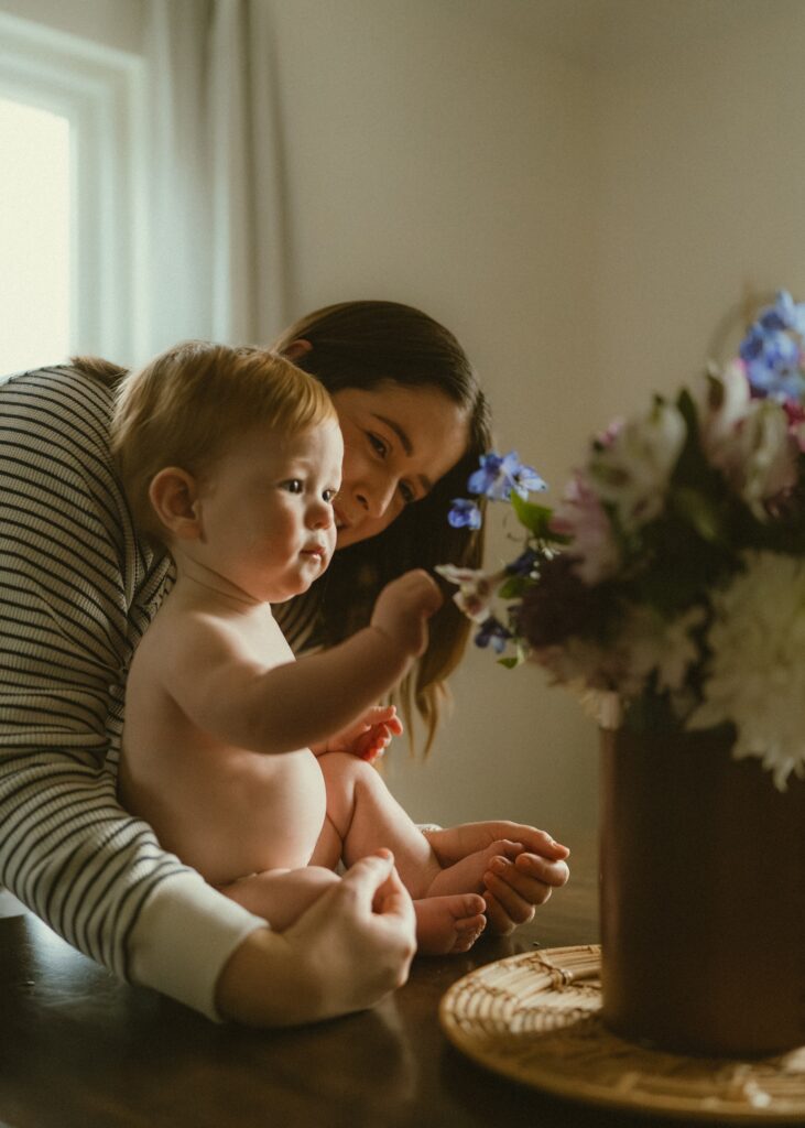 Mom leaning over her toddler son while he picks at flowers on the dining room table