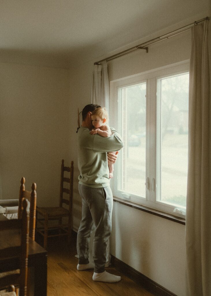 Dad holding toddler son looking outside the window in their home during their comfortable in home photo session in Cincinnati, Ohio