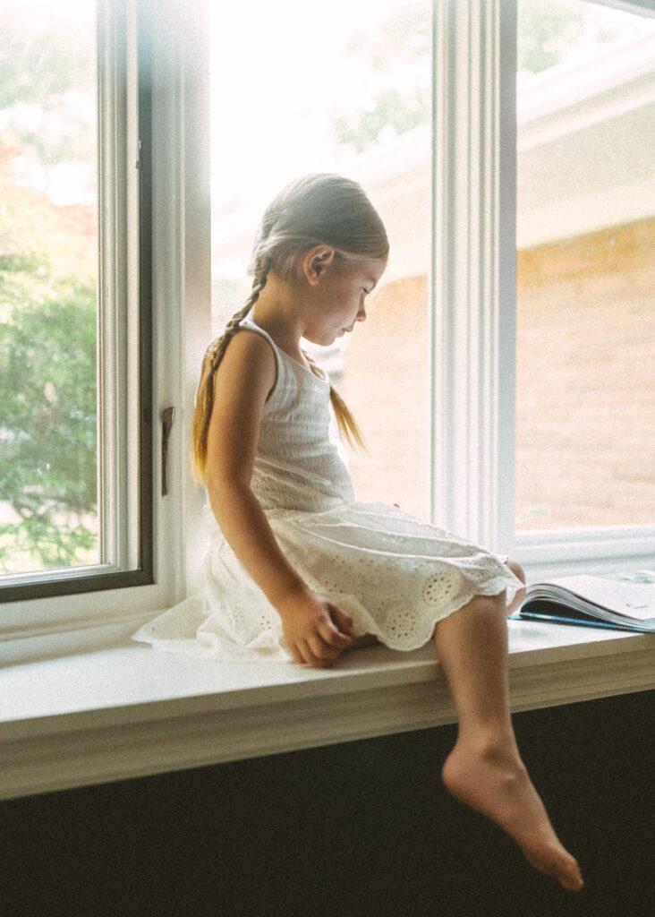 toddler girl wearing a white sleeveless dress sitting in the window sill with her book