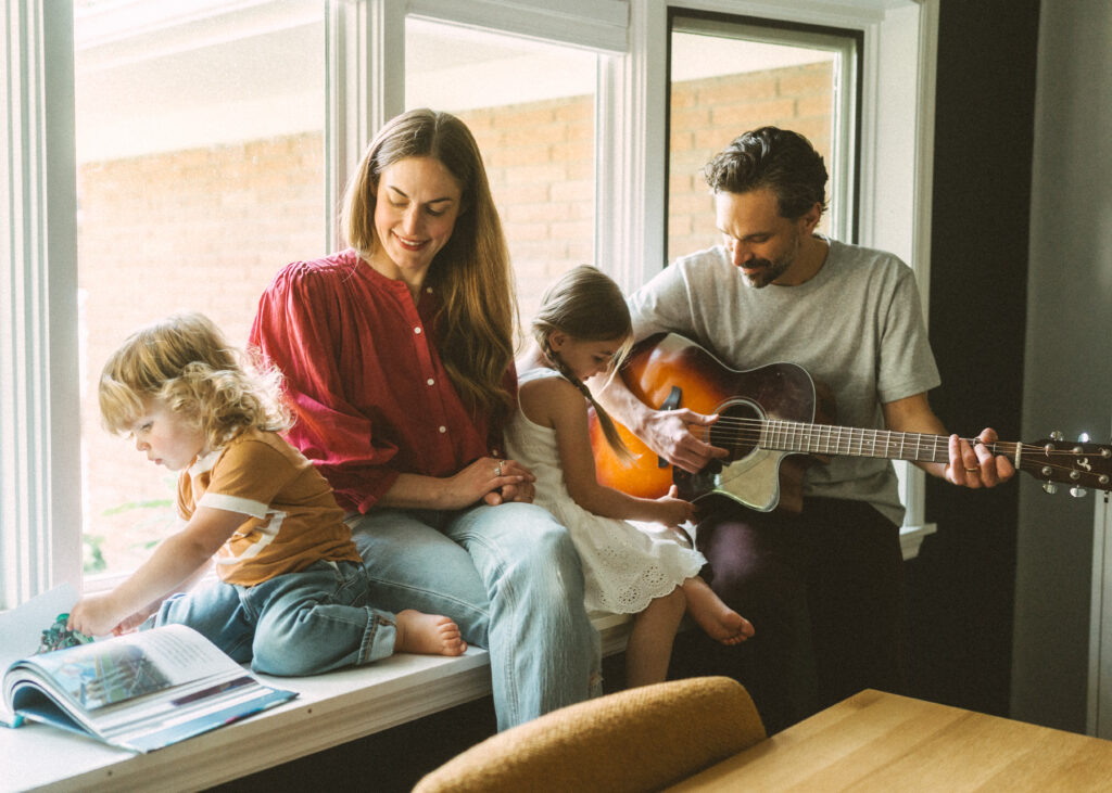 mom helping toddler boy read and dad playing the guitar for his toddler daughter while all are sitting in their large window sill.