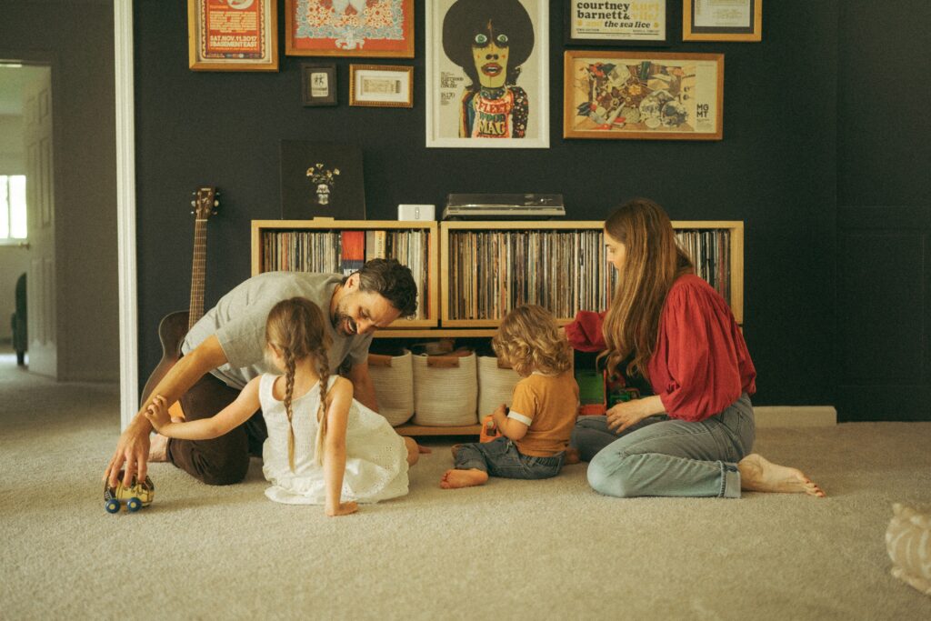 a mom and dad sitting on the floor with their toddler girl and boy playing with toy cars sitting in front of their record collection