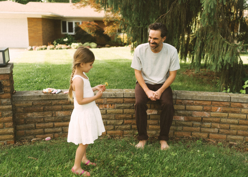 dad sitting on a brick retaining wall in front of their home smiling at his toddler daughter holding a dandelion