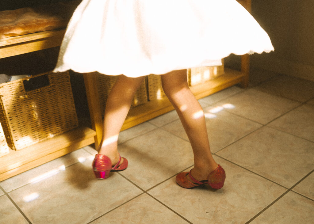toddler girl wearing white dress and pink heels dancing in dappled light showing close up of her shoes and legs