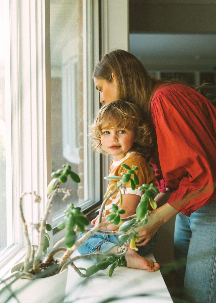 Toddler boy sitting in window sill being comforted by his mom near plants
