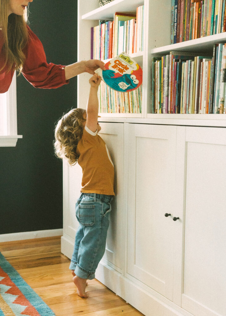 Toddler boy in jeans and orange shirt reaching for children's book off of shelf standing on his toes