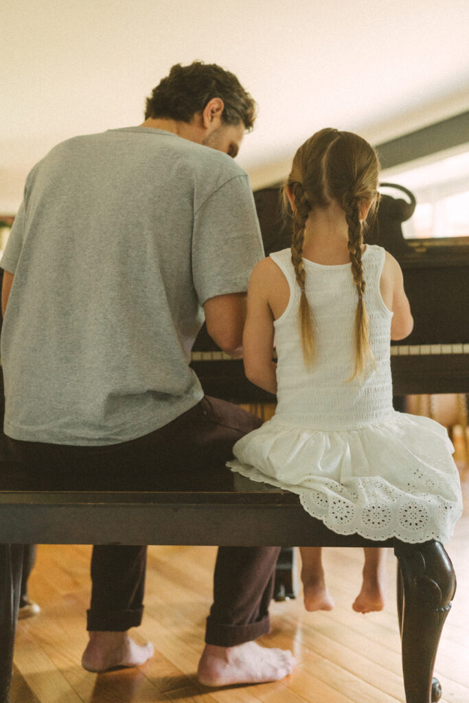 back shot of a father showing his toddler girl in pigtail braids how to play the piano