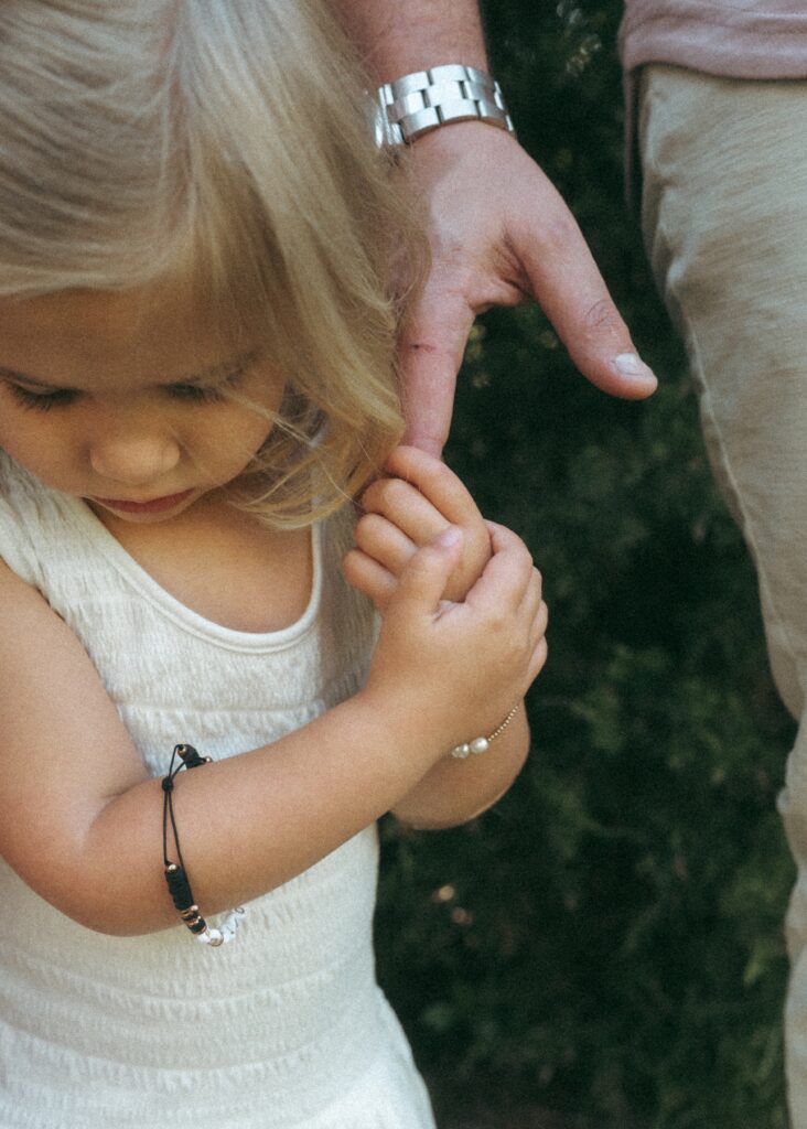 Close up image of blonde toddler girl in a white dress holding her dad's finger