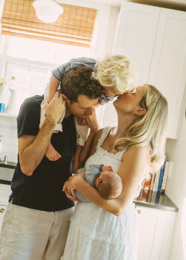 family of four in their kitchen with toddler on dad's shoulders and leaning down to kiss mom